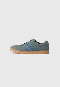 Sneaker de ante verde claro con acentos azules, cordones planos y una suela de goma beige. Punta redondeada y diseño de perfil bajo.