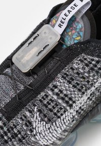 Black and gray knit sneaker featuring a textured design, a clear upper clip, and a colorful patterned inner lining. Transparent sole accents.