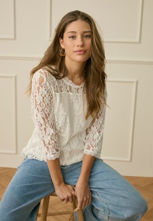 Jauna moteris su ilgais plaukais, vilkinti balta nėriniuota palaidine ir mėlynais džinsais, sėdi ant medinio kėdės priešais smėlio spalvos lentelėmis apkaltą sieną.