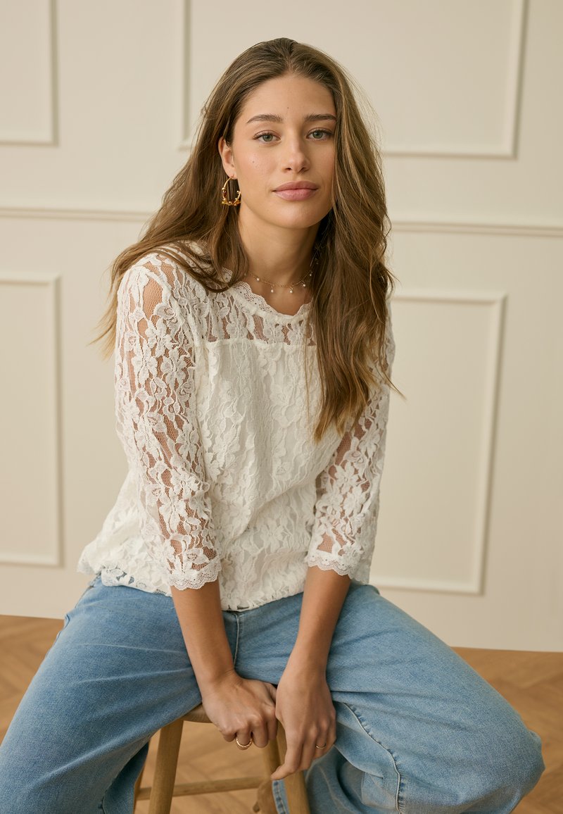 Mlada ženska z dolgimi lasmi, oblečena v belo čipkasto majico in modre kavbojke, sedi na lesenem stolu ob bež obzidju z lesenimi paneji.