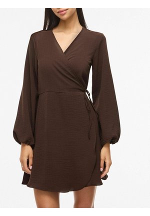 Femme portant une robe portefeuille marron à manches longues avec un col en V et un détail de nouage à la taille, debout devant un fond blanc uni.