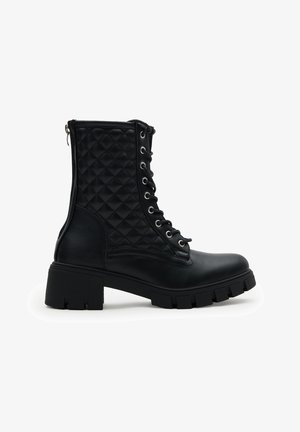 Botas de combate de cuero negro con paneles laterales acolchados, parte delantera con cordones, suela gruesa y cierre de cremallera en la parte trasera, mostradas en perfil lateral.