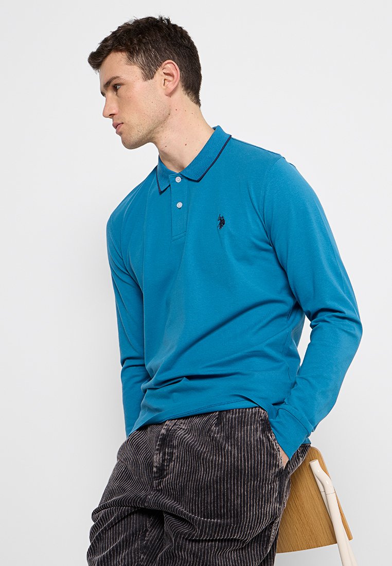 Polo a maniche lunghe in cotone blu brillante, con colletto e logo ricamato nero. Abbinato a pantaloni in velluto a coste scuri a righe.