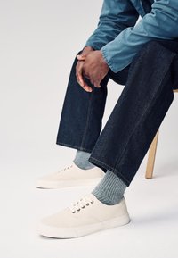 Smėlio spalvos drobiniai sportbačiai su baltais guminiais padais, derinami su pilkomis riboto audinio kojinėmis ir tamsiomis džinsinėmis kelnėmis. Minimalistinis dizainas su skylutėmis ir batraišteliais.