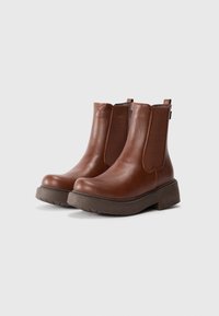 Botas de cuero marrón hasta el tobillo con paneles laterales elásticos, puntera redonda y suela gruesa gris; textura suave y detalles de costura mínimos.