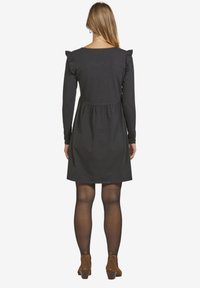 Schwarzes Kleid mit langen Ärmeln, gerafften Schulterakzenten und einem taillierten Oberteil. Ausgestellter Rock reicht bis zur Mitte des Oberschenkels und wird mit braunen Knöchelstiefeln kombiniert.