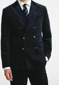 Man i en mörk dubbelknäppt snickeri kostym med en vit skjorta och marint randig slips, med en hand i fickan, mot en enfärgad bakgrund.