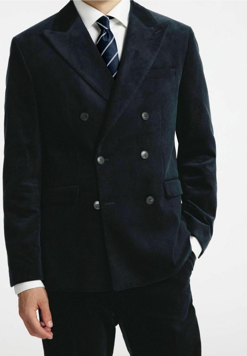 Man i en mörk dubbelknäppt snickeri kostym med en vit skjorta och marint randig slips, med en hand i fickan, mot en enfärgad bakgrund.