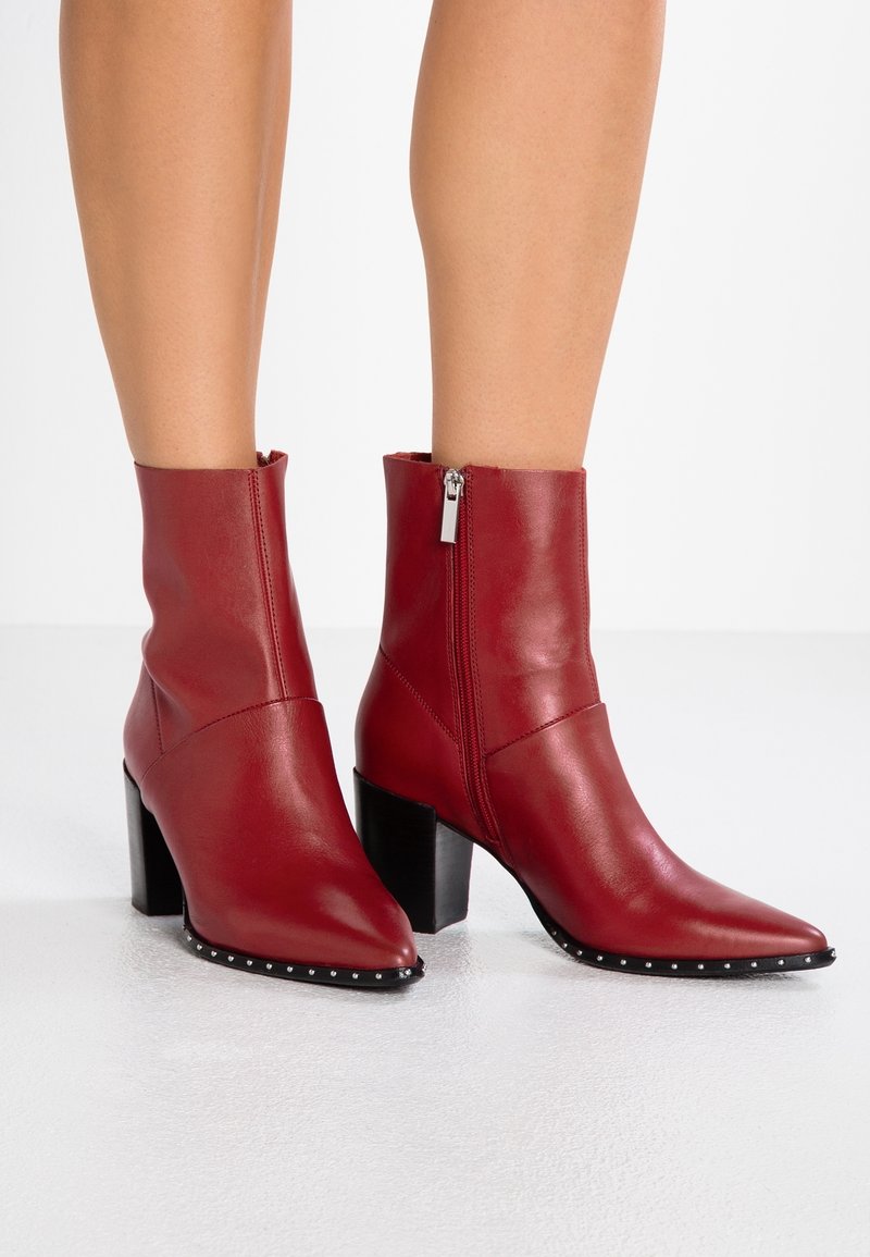 Bottes en cuir rouge montant à la cheville, avec bouts pointus, fermetures éclair latérales, bords cloutés et talons blocs noirs, portées sur jambes nues.