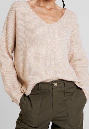 Persona con un suéter de punto beige con cuello en V y pantalones verde oliva con las manos en los bolsillos.