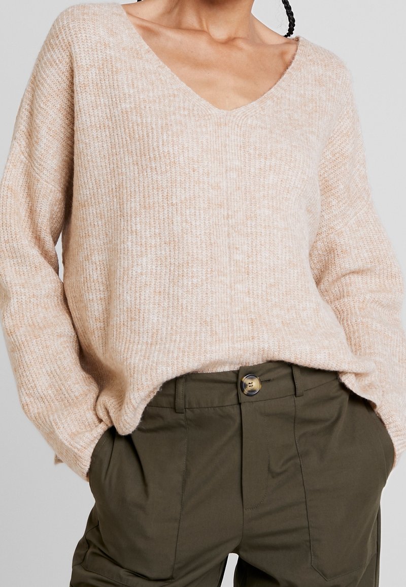 Personne portant un pull beige en tricot avec un col en V et un pantalon vert olive, les mains dans les poches.