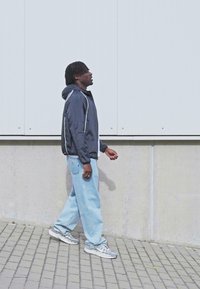 Sportjakke i marineblå med hvide accenter, løse lyseblå jeans og grå sneakers. Personen går langs et struktureret stenbelægning.