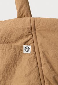 Ruskea kangaskassi, jossa on ommeltu tasku ja valkoinen etiketti mustalla pyöreällä monogrammilogolla.