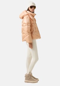 Brzoskwiniowa kurtka puchowa z kapturem, w połączeniu z białymi legginsami i beżowymi butami wyłożonymi futrem. Okulary przeciwsłoneczne i biała dziana czapka dopełniają stylizację.