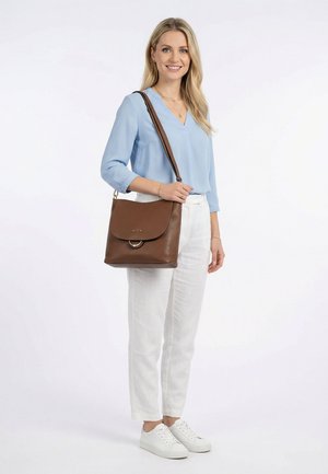 Femme portant un chemisier bleu clair, un pantalon blanc et des baskets blanches, portant un grand sac à épaule en cuir marron, souriant devant un fond blanc.