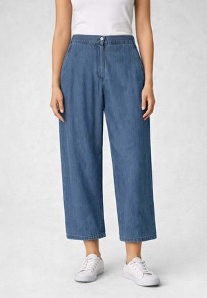 Donna che indossa jeans blu a vita alta, gamba larga e accorciati, con bottone frontale, abbinati a sneakers bianche e top bianco senza maniche.