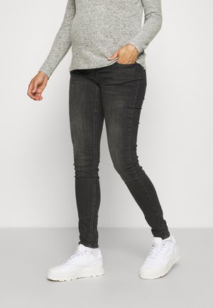 Personne portant un jean skinny gris foncé, un pull à manches longues gris clair et des baskets blanches, debout avec une main dans la poche.