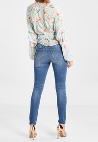 Kukkakuvioinen pusero, jossa on röyhelöhiukset, yhdistettynä kapeisiin sinisiin farkkuihin. Mallilla on yllään kiiltävät nude-korkokengät.