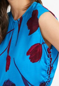 Mouwloos topje van levendig blauw stof, met een bloemenpatroon in donkerrood en paars. Gerepte details bij de armsgaten voegen textuur toe.