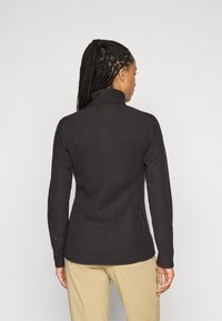 Donna con capelli intrecciati, che indossa una giacca nera aderente e testurizzata e pantaloni beige, vista da dietro su uno sfondo semplice.