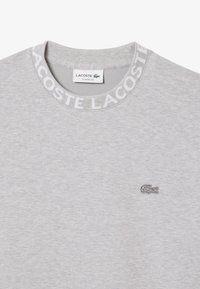 Sweatshirt gris à col rond, avec un col texturé portant "LACOSTE" en blanc. Inclut un petit logo crocodile brodé.