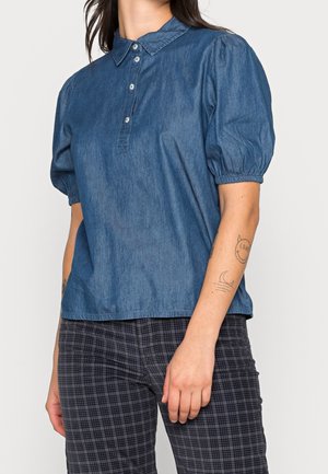 Kobieta w niebieskiej denimowej bluzce z krótkimi, bufkami rękawami oraz ciemnych, kratowanych spodniach, pokazująca tatuaże na lewym przedramieniu.