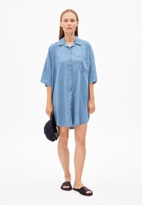 Lichtblauwe denim jurk met korte mouwen, knopsluiting aan de voorkant, een borstzak en een ontspannen pasvorm. Gemaakt met zwarte slide sandalen.