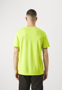 Hombre con cabello corto y tatuajes en los brazos, vistiendo una camiseta amarilla de manga corta y pantalones oscuros, de pie de espaldas sobre un fondo blanco.