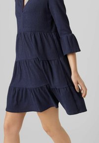 Navyblaues Kleid aus strukturiertem Stoff, mit drei gestuften Lagen und Schlagärmel. Der V-Ausschnitt verleiht der Silhouette Eleganz.