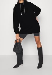 Mujer vestida con una sudadera negra oversized, pantalones cortos de ciclista grises, botas altas negras de tacón, sosteniendo un bolso de malla negro frente a un fondo blanco.