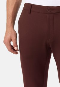 Nahaufnahme einer linken Hand neben bordeauxfarbenen, geknöpften Hosen mit Gürtelschlaufen vor einem einfachen hellen Hintergrund.