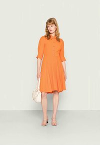 Vestido laranja com botões, saia rodada, mangas curtas bufantes e um tecido texturizado. Combinado com uma mala branca e sandálias.