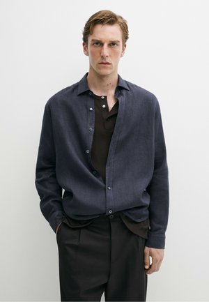 Jeune homme aux cheveux châtain clair portant une chemise bleu foncé boutonnée sur un henley noir, les mains dans les poches d'un pantalon noir, debout devant un fond blanc.