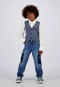 Μπλε γιλέκο με καρώ πάνω από λευκό πουκάμισο, συνδυασμένο με σκισμένο μπλε τζιν με σκούρες κηλίδες. Μαύρα και λευκά αθλητικά παπούτσια ολοκληρώνουν την εμφάνιση.