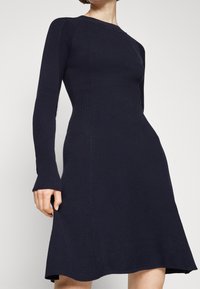Granatowa sukienka z prążkowanego dzianego materiału, z długimi rękawami, dekoltem w łódkę i rozkloszowanym dołem. Kontrast faktur pomiędzy gładkimi a prążkowanymi partiami.