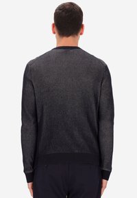 Uomo che indossa un maglione a coste scuro a maniche lunghe e pantaloni scuri, in piedi con la schiena rivolta verso uno sfondo bianco semplice.