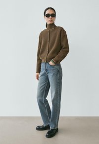 Brauner Rippenstrick-Rollkragenpullover mit Knöpfen, kombiniert mit hellblauen Denim-Jeans und schwarzen klobigen Schuhen, stehend vor einem neutralen Hintergrund.