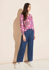 Long-sleeve pink patterned blouse with floral design, paired with wide-leg blue denim pants and white sneakers. Smooth fabric texture.