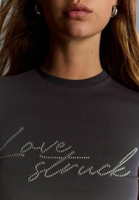 Kvinna som bär en mörk topp med silvernitar och kursiv text "Lovely Sweet," där hon visar sin nacke, läppar och ringörhängen.