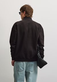 Mand med kort brunt hår iført sort jakke og lyseblå jeans, der holder en lille sort taske, vendt væk mod en ensfarvet baggrund.