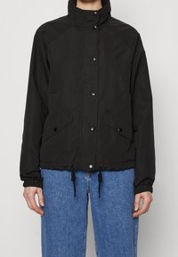 Svart knappad jacka med hög krage, fickor med klaff, dragsko i nederkant, bärs med blå jeans.