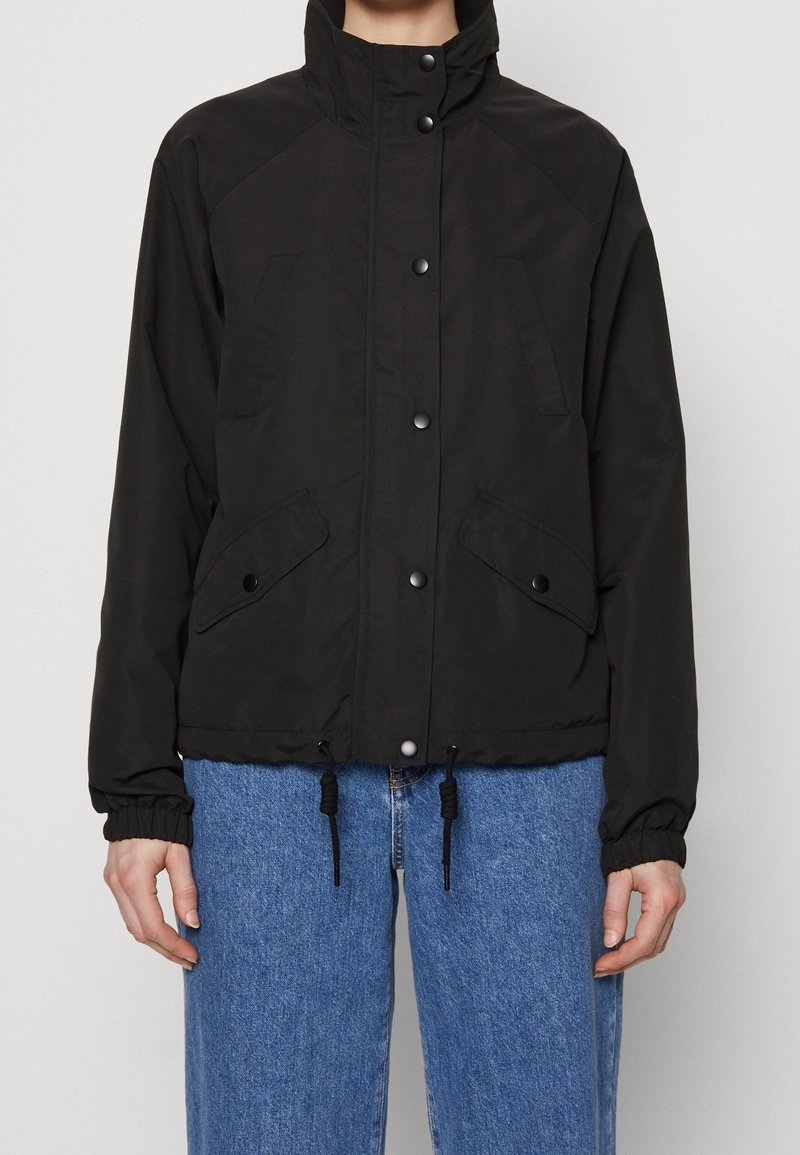 Svart knappad jacka med hög krage, fickor med klaff, dragsko i nederkant, bärs med blå jeans.