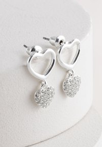 Pendientes de plata con diseños en forma de corazón. El corazón superior es abierto, con un corazón colgante adornado con pequeños cristales transparentes.