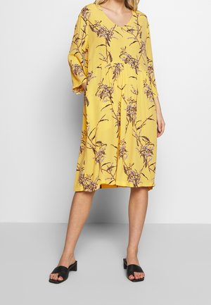 Žluté květinové šaty s výstřihem do V, tříčtvrtečními rukávy a volným střihem. Vzor tvoří rozkvetlá větev; nošeny s černými pantoflemi.