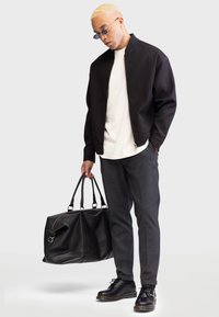Svart bomberjakke med teksturert stoff, hvit t-skjorte, grå bukser, svarte lærsko og en svart duffelbag med metalldetaljer.