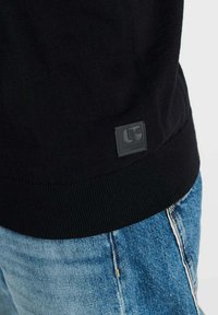 Zwarte trui met geribbelde zoom, voorzien van een textuurlabel met "LC"-logo. Gecombineerd met lichtblauwe spijkerbroek met vervaagde details.