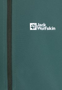 Grön tyg med en texturerad yta, en vertikal svart dragkedja och en vit logotyp med ett tassavtryck och texten "Jack Wolfskin."