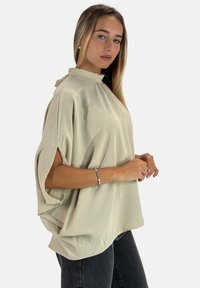 Hellbeige oversized Bluse aus strukturiertem Stoff, mit hohem Kragen und weiten Ärmeln, kombiniert mit dunklen Jeans. Einfaches und minimalistisches Design.