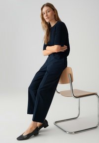 Woman with long blonde hair in black knit top and navy pants sitting on the edge of a modern wooden chair with metal frame.