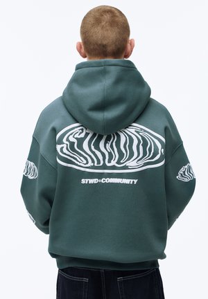 Persona che indossa una felpa con cappuccio color verde acqua con un audace motivo astratto bianco e il testo "STWD COMMUNITY" sul retro, rivolta di spalle alla fotocamera.